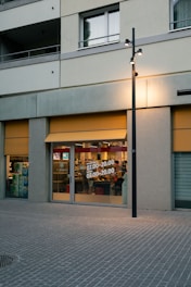 A storefront on a street with large glass windows displaying store hours. The exterior has a gray facade with yellow awnings. Inside, various store items are visible, and there is a reflection of interior lighting. A streetlight is located in front of the store.