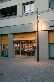 A storefront on a street with large glass windows displaying store hours. The exterior has a gray facade with yellow awnings. Inside, various store items are visible, and there is a reflection of interior lighting. A streetlight is located in front of the store.