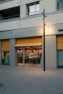 A storefront on a street with large glass windows displaying store hours. The exterior has a gray facade with yellow awnings. Inside, various store items are visible, and there is a reflection of interior lighting. A streetlight is located in front of the store.