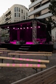 An outdoor stage is set up with bright pink lighting against the backdrop of modern apartment buildings. Wooden benches are arranged in front of the stage, suggesting a venue prepared for an event or performance.