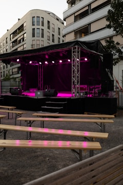 An outdoor stage is set up with bright pink lighting against the backdrop of modern apartment buildings. Wooden benches are arranged in front of the stage, suggesting a venue prepared for an event or performance.