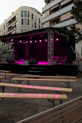 An outdoor stage is set up with bright pink lighting against the backdrop of modern apartment buildings. Wooden benches are arranged in front of the stage, suggesting a venue prepared for an event or performance.