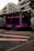 An outdoor stage is set up with bright pink lighting against the backdrop of modern apartment buildings. Wooden benches are arranged in front of the stage, suggesting a venue prepared for an event or performance.