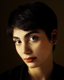 A portrait of a person with short dark hair and prominent facial features, including large eyes and full lips. The lighting highlights their face against a dark background, creating a dramatic effect.