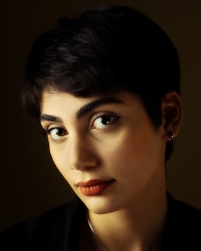 A portrait of a person with short dark hair and prominent facial features, including large eyes and full lips. The lighting highlights their face against a dark background, creating a dramatic effect.
