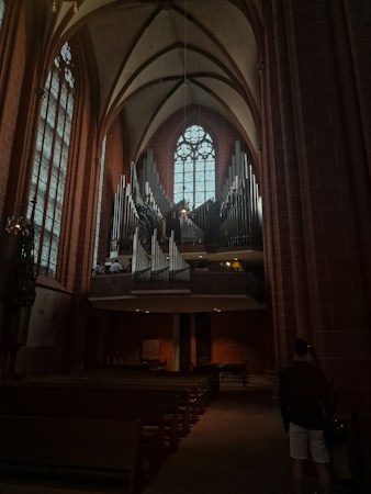 A large pipe organ is prominently displayed in a spacious and dimly lit church interior. The ceiling is vaulted, with arched beams and tall stained glass windows lining the walls. Rows of wooden pews are visible, and a couple of people are seen near the organ, providing a sense of scale.