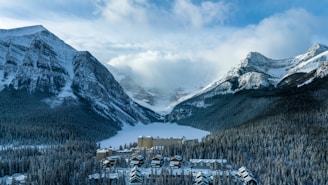 A snow-covered mountain landscape features a large lake frozen in the middle, surrounded by dense forests. Nestled at the foot of the mountains near the lake is a grand hotel with multiple buildings showing alpine architecture.
