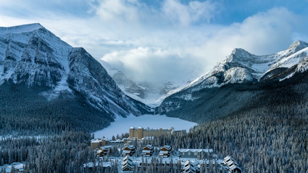 A snow-covered mountain landscape features a large lake frozen in the middle, surrounded by dense forests. Nestled at the foot of the mountains near the lake is a grand hotel with multiple buildings showing alpine architecture.
