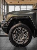 The image features a close-up of the front section of a rugged, modern SUV or truck parked inside a showroom. The focus is on the large, intricately designed wheel with prominent tread patterns and stylish alloy rims. The body of the vehicle is painted in a matte green or gray color, with sleek lines and a robust aesthetic. Large windows and polished tiles compose the backdrop, illuminating the vehicle's surface with natural and artificial light.