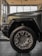 The image features a close-up of the front section of a rugged, modern SUV or truck parked inside a showroom. The focus is on the large, intricately designed wheel with prominent tread patterns and stylish alloy rims. The body of the vehicle is painted in a matte green or gray color, with sleek lines and a robust aesthetic. Large windows and polished tiles compose the backdrop, illuminating the vehicle's surface with natural and artificial light.
