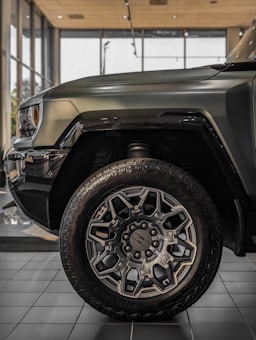 The image features a close-up of the front section of a rugged, modern SUV or truck parked inside a showroom. The focus is on the large, intricately designed wheel with prominent tread patterns and stylish alloy rims. The body of the vehicle is painted in a matte green or gray color, with sleek lines and a robust aesthetic. Large windows and polished tiles compose the backdrop, illuminating the vehicle's surface with natural and artificial light.