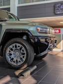A close-up view of a rugged, modern SUV with large, intricate alloy wheels. The vehicle is parked indoors on a tiled floor with a partially unrolled mat in front of it. The setting appears to be a dealership with bright lighting and a clean, polished environment.