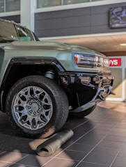 A close-up view of a rugged, modern SUV with large, intricate alloy wheels. The vehicle is parked indoors on a tiled floor with a partially unrolled mat in front of it. The setting appears to be a dealership with bright lighting and a clean, polished environment.