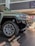A close-up view of a rugged, modern SUV with large, intricate alloy wheels. The vehicle is parked indoors on a tiled floor with a partially unrolled mat in front of it. The setting appears to be a dealership with bright lighting and a clean, polished environment.