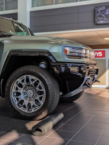 A close-up view of a rugged, modern SUV with large, intricate alloy wheels. The vehicle is parked indoors on a tiled floor with a partially unrolled mat in front of it. The setting appears to be a dealership with bright lighting and a clean, polished environment.