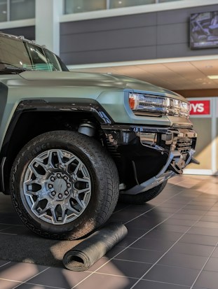 A close-up view of a rugged, modern SUV with large, intricate alloy wheels. The vehicle is parked indoors on a tiled floor with a partially unrolled mat in front of it. The setting appears to be a dealership with bright lighting and a clean, polished environment.