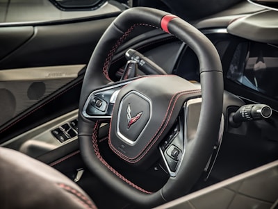 A detailed view of a car's steering wheel adorned with black leather and red stitching, featuring a logo in the center. Control buttons are visible on the spokes, alongside a turn signal lever. The surrounding interior is sleek, with a visible Bose speaker and window controls integrated into the door panel.