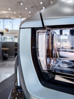 Close-up view of a car headlight with a modern design, featuring sleek lines and reflective surfaces. The headlights are set within a metallic frame, indicating a robust and stylish automotive aesthetic. Background shows an indoor car showroom with blurred details and ceiling lights.