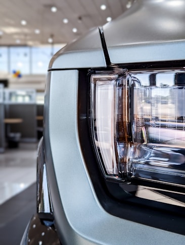 Close-up view of a car headlight with a modern design, featuring sleek lines and reflective surfaces. The headlights are set within a metallic frame, indicating a robust and stylish automotive aesthetic. Background shows an indoor car showroom with blurred details and ceiling lights.