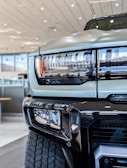 Close-up view of the front section of a truck, highlighting the headlight design and part of the grille. The vehicle is parked indoors, likely in a showroom, with modern architecture visible in the background. The tire tread is prominently visible in the lower part of the image.