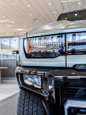 Close-up view of the front section of a truck, highlighting the headlight design and part of the grille. The vehicle is parked indoors, likely in a showroom, with modern architecture visible in the background. The tire tread is prominently visible in the lower part of the image.