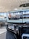 Close-up view of the front section of a truck, highlighting the headlight design and part of the grille. The vehicle is parked indoors, likely in a showroom, with modern architecture visible in the background. The tire tread is prominently visible in the lower part of the image.