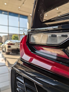 Close-up view of a car's tail light with a modern design, featuring sleek lines and a combination of red and black colors. In the background, another car can be seen parked in a showroom with large glass windows, allowing natural light to illuminate the space.