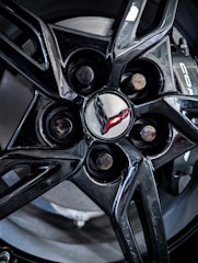 A close-up view of a shiny black alloy wheel with a branded logo at its center. The logo consists of a stylized flag emblem, partially in red, set against a metallic gray center cap.