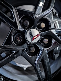 A close-up view of a shiny black alloy wheel with a branded logo at its center. The logo consists of a stylized flag emblem, partially in red, set against a metallic gray center cap.