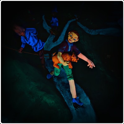 A vividly colored figurine of a person with exaggerated facial features is holding an orange cat while standing on a narrow stream. Another figure in a blue outfit is seen in the background, partially shadowed.