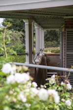 A covered porch with white pillars supports a corrugated metal roof. The railing runs along the edge, and there are lush green plants and white flowers in the foreground. Brick walls form part of the structure, and there are shutters on the windows.