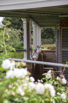 A covered porch with white pillars supports a corrugated metal roof. The railing runs along the edge, and there are lush green plants and white flowers in the foreground. Brick walls form part of the structure, and there are shutters on the windows.