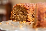 A close-up view of a slice of homemade cake with a moist and crumbly texture. The cake is brown, indicating it might be made with ingredients like cinnamon or brown sugar, and there are visible nuts embedded within the slice.