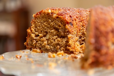 A close-up view of a slice of homemade cake with a moist and crumbly texture. The cake is brown, indicating it might be made with ingredients like cinnamon or brown sugar, and there are visible nuts embedded within the slice.