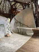A modern, organic interior space featuring curved, smooth surfaces and natural elements. The ceiling is made of woven wooden branches, creating a rustic texture. A sleek, built-in bench curves along the wall, which is adorned with a wooden branch and small climbing plant. There are potted plants placed on the floor, and the overall aesthetic is both contemporary and earthy.