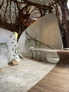 A modern, organic interior space featuring curved, smooth surfaces and natural elements. The ceiling is made of woven wooden branches, creating a rustic texture. A sleek, built-in bench curves along the wall, which is adorned with a wooden branch and small climbing plant. There are potted plants placed on the floor, and the overall aesthetic is both contemporary and earthy.