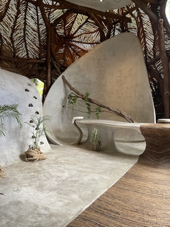 A modern, organic interior space featuring curved, smooth surfaces and natural elements. The ceiling is made of woven wooden branches, creating a rustic texture. A sleek, built-in bench curves along the wall, which is adorned with a wooden branch and small climbing plant. There are potted plants placed on the floor, and the overall aesthetic is both contemporary and earthy.