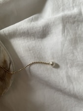 A delicate gold chain with small beads, adorned with a single pearl, is elegantly placed atop a soft, textured white fabric. The light casts gentle shadows, creating a serene and minimalist atmosphere.
