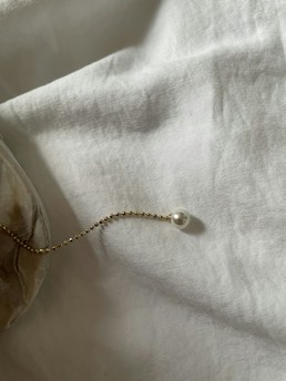A delicate gold chain with small beads, adorned with a single pearl, is elegantly placed atop a soft, textured white fabric. The light casts gentle shadows, creating a serene and minimalist atmosphere.