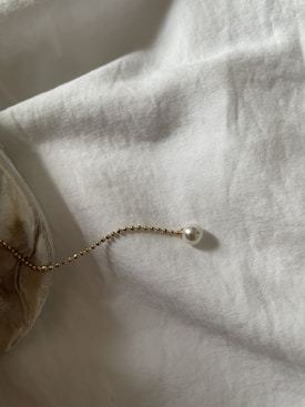 A delicate gold chain with small beads, adorned with a single pearl, is elegantly placed atop a soft, textured white fabric. The light casts gentle shadows, creating a serene and minimalist atmosphere.