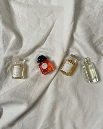 Four small glass perfume bottles of various shapes and colors are laid out on a soft, white fabric. The bottles are evenly spaced and include scents labeled as 'Replica', 'Twilly', and 'Hermes'. The gentle folds of the fabric create a simple, elegant backdrop.