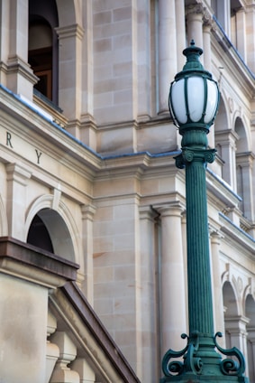 A detailed architectural view featuring a historic beige stone building with classical pillars and arches. In the foreground, an ornate vintage green lamp post is present, adding a decorative element.