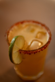 A cocktail glass filled with an iced drink is garnished with a slice of lime. The rim of the glass is coated in a red spice, likely chili powder or similar seasoning.