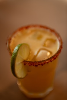 A cocktail glass filled with an iced drink is garnished with a slice of lime. The rim of the glass is coated in a red spice, likely chili powder or similar seasoning.