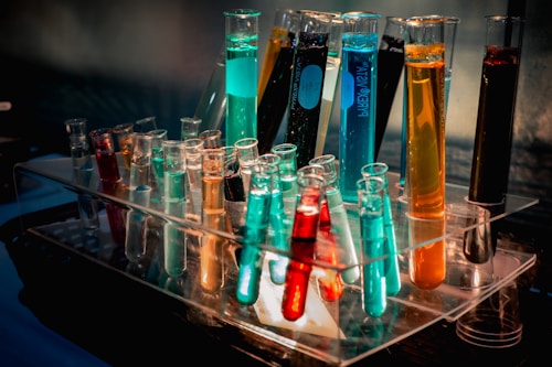 Several test tubes filled with various colored liquids are arranged in a clear test tube rack. The liquids display a range of vibrant colors including orange, blue, turquoise, and red.