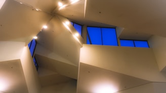 Complex, angular architecture with sharp lines and geometric shapes features prominently, with multiple recessed lighting fixtures and large windows letting in evening light.