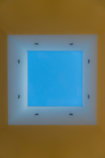 A square skylight is centered in a ceiling, surrounded by recessed lighting fixtures. The view through the skylight reveals a serene blue sky.