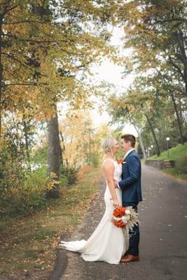A couple, dressed in wedding attire, stands closely together on a tree-lined pathway. The bride is wearing a white dress and holding a bouquet of vivid orange flowers. The groom is in a blue suit, complementing the autumnal setting with leaves in yellow and orange hues.