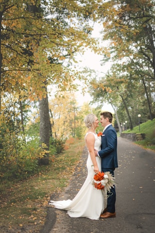 A couple, dressed in wedding attire, stands closely together on a tree-lined pathway. The bride is wearing a white dress and holding a bouquet of vivid orange flowers. The groom is in a blue suit, complementing the autumnal setting with leaves in yellow and orange hues.