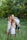 A family gathers together in a lush green outdoor setting, with grass underfoot and leafy trees in the background. The group consists of two adults and four children. The children are dressed in light summer clothing, and one child is being held by an adult.
